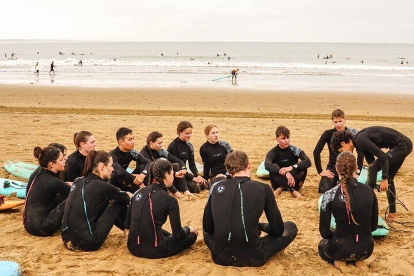 Un groupe de personnes vêtues de combinaisons de plongée est assis sur la plage, certaines tenant des planches de surf. 3