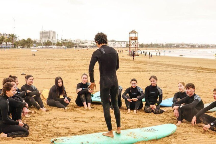 Un groupe de personnes en combinaisons de plongée sur une plage de sable, avec des planches de surf, une tour de surveillance et des bâtiments en arrière-plan. 4