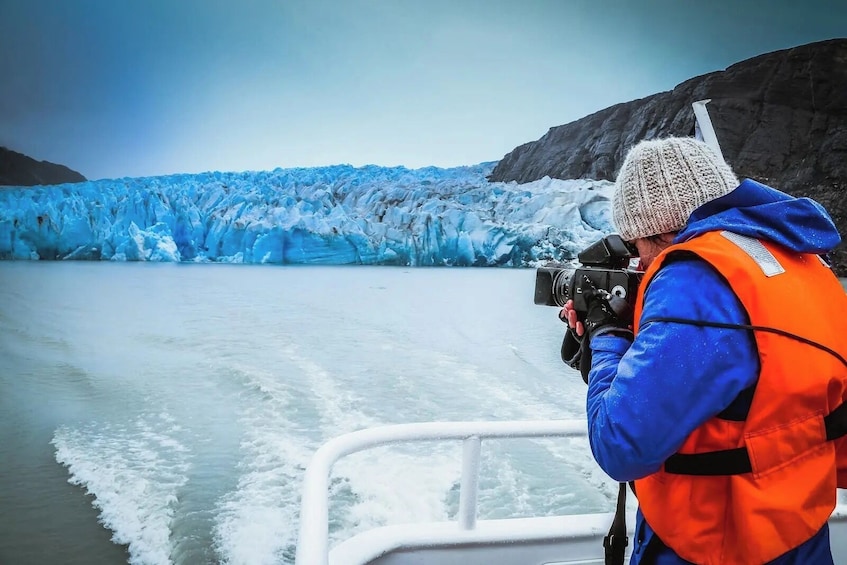 A person in a blue jacket and orange vest taking a photo of a glacier. 2