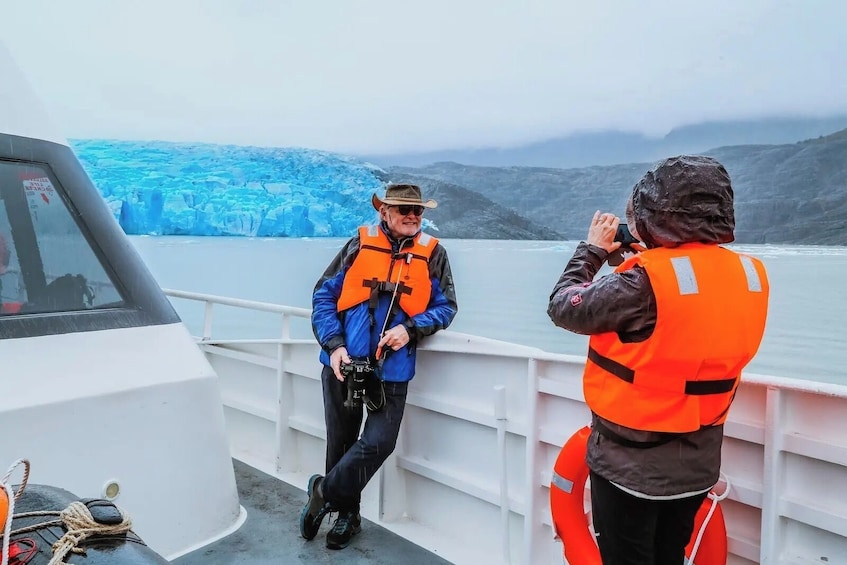 Two people on a boat wearing life jackets, one holding a camera, with a glacier in the background. 3