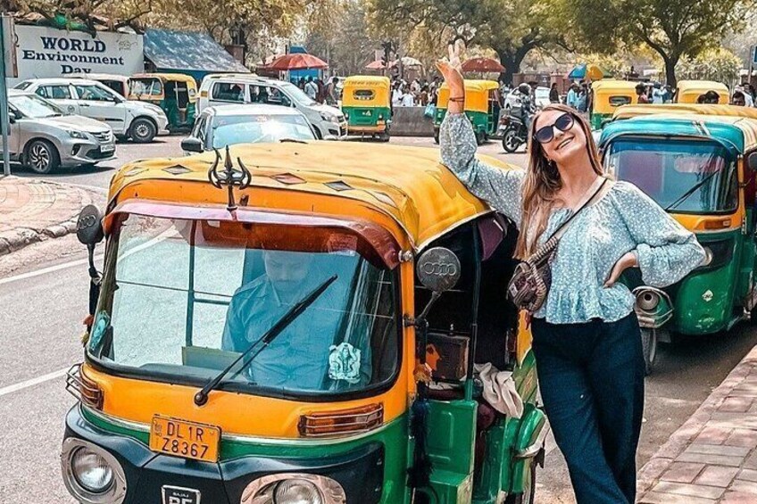 Une personne se tient à côté d’un auto-rickshaw jaune et vert, portant une plaque d’immatriculation, dans un environnement urbain où l’on aperçoit d’autres véhicules et une affiche pour la Journée mondiale de l’environnement. 1