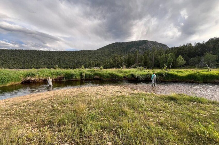 A riverbank with two people fishing, surrounded by greenery and a forested hill. 2