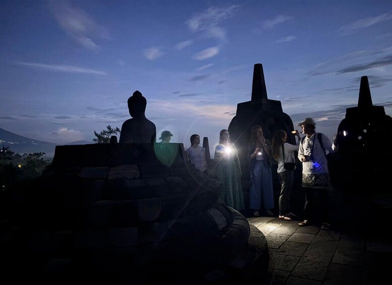 A group of people standing around a large, dark structure at dusk with a mountain in the background. 2