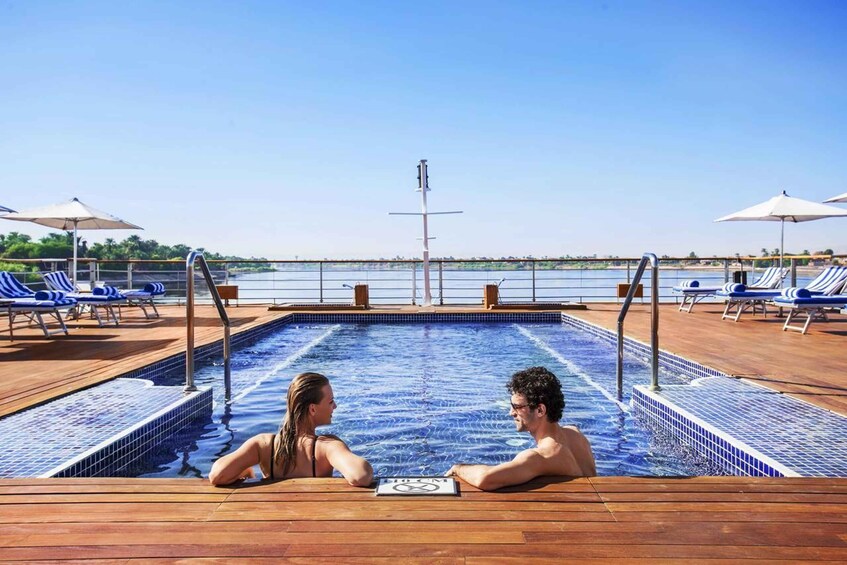 A couple in a hot tub on a deck with a pool and lounge chairs, overlooking a body of water. 3