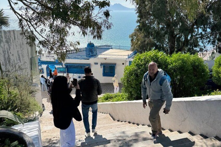 Una cittadina costiera con persone che salgono le scale, un edificio a cupola blu e vista sul mare. 2