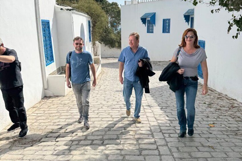 Tre persone camminano su un sentiero acciottolato tra edifici bianchi con dettagli blu. 5