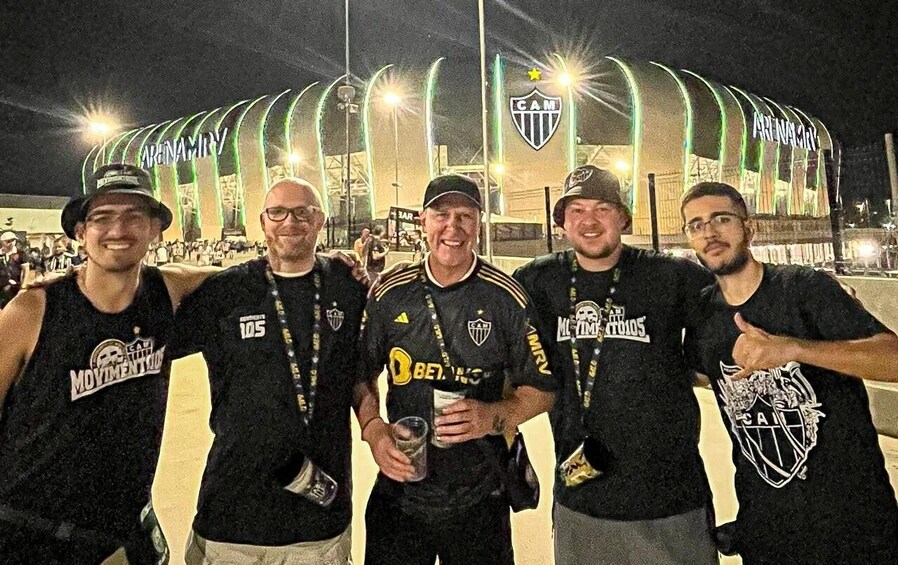 A group of people in black sports jerseys with logos, standing in front of a stadium with a distinctive arch and the letters 'CAM' on it. 1