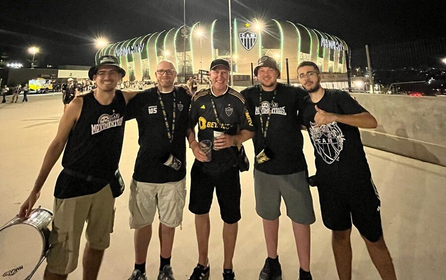 Five people standing in front of a stadium with a large arch and a star logo. 5