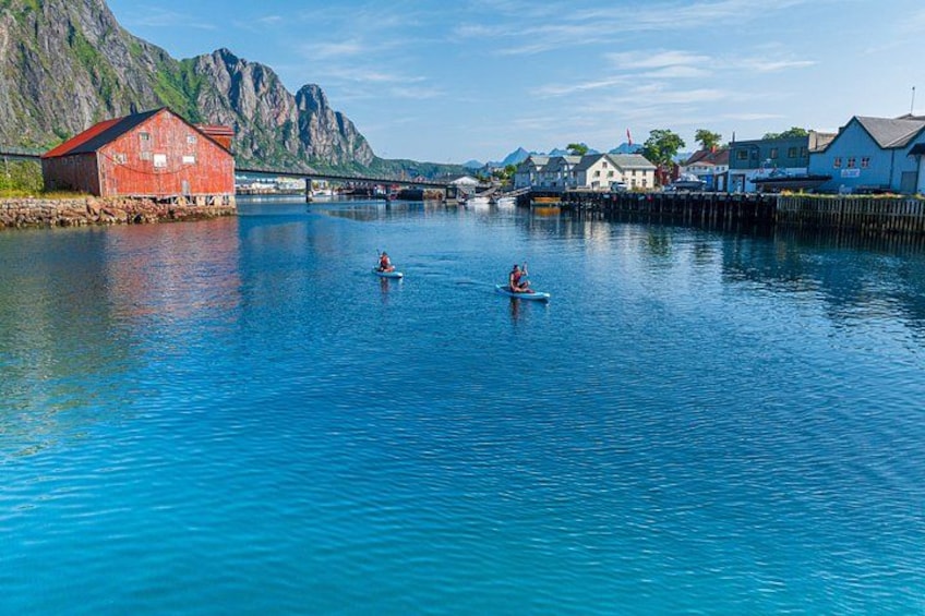 A calm river with kayakers, a red building, and a bridge in the background. 1