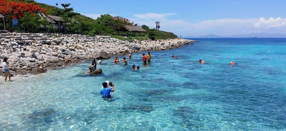 A beach with clear water, people swimming and sunbathing, and a rocky shoreline. 2