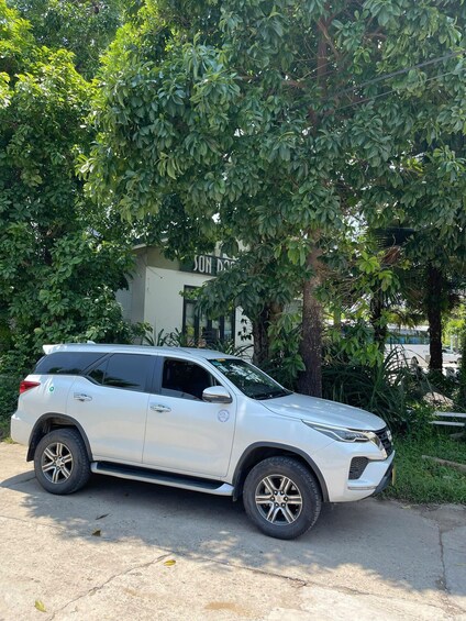 A white SUV parked under a tree with a building labeled 'SON DOY' in the background. 2