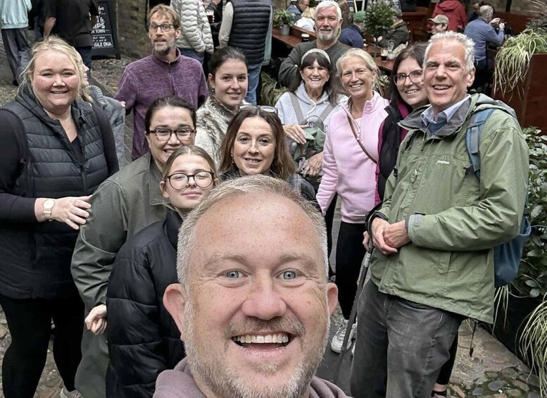 A group of people gathered outdoors, possibly at a public event or market, with stone pavement and greenery in the background. 1