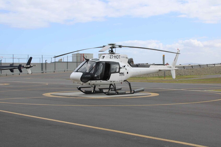 A white helicopter with 'ZT-HOT' and 'LICOPTERS' on its side parked on a tarmac with yellow lines and a helipad symbol. 1