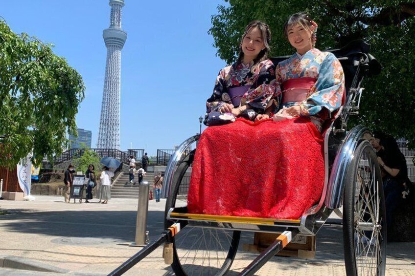 Twee personen in traditionele Japanse kledij zitten in een rijksja, met een moderne toren op de achtergrond. 1
