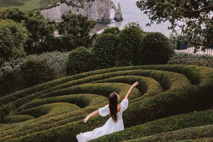 Una mujer con un vestido blanco de pie en un jardín con un laberinto, una gran formación rocosa y un cuerpo de agua al fondo. 1