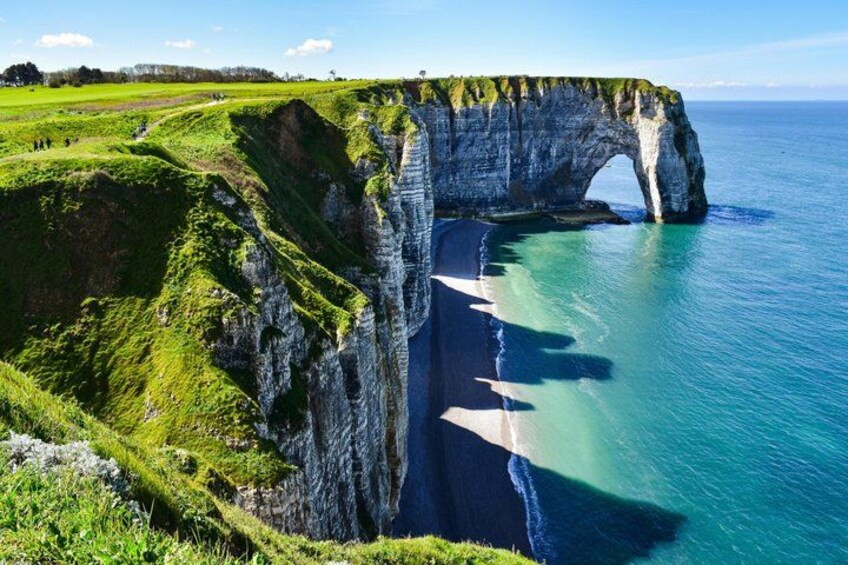 A coastal cliff with a natural arch formation, a beach, and a grassy landscape. 1