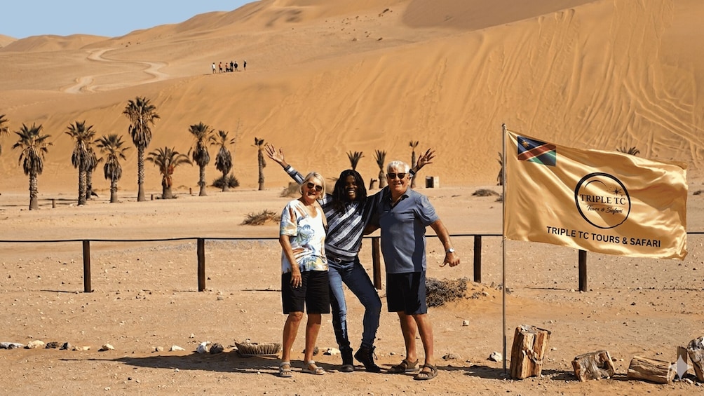 Three people posing with a flag in a desert landscape with sand dunes and palm trees. 3