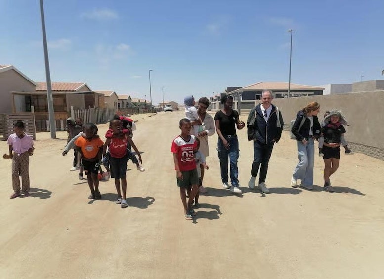 Eine Gruppe von Menschen geht auf einem Sandweg, im Hintergrund sind Gebäude und ein klarer Himmel zu sehen. 2