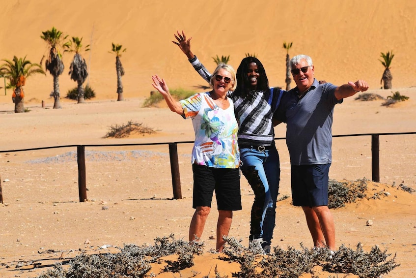 Three people standing in a desert with palm trees and a fence in the background. 5