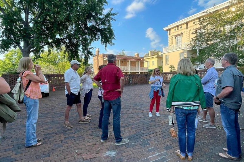 A group of people standing on a brick pavement, some facing away from the camera, in an outdoor setting with trees and buildings in the background. 2