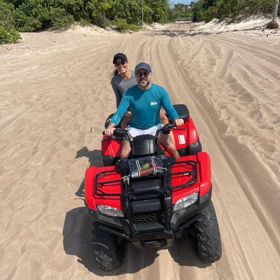 Two people on a red ATV in a sandy area with sparse vegetation. 1