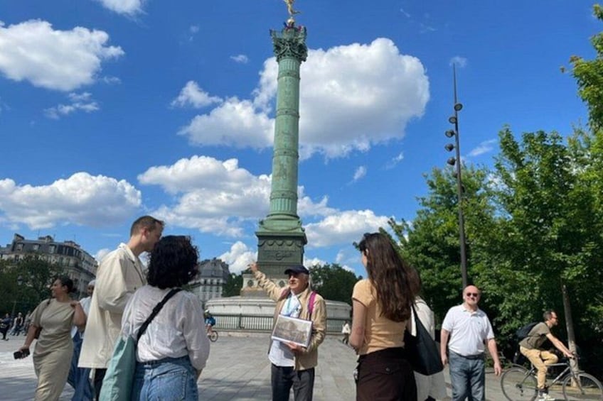 A group of people standing around a tall green monument with a statue on top, in an open square with trees and buildings in the background. 1