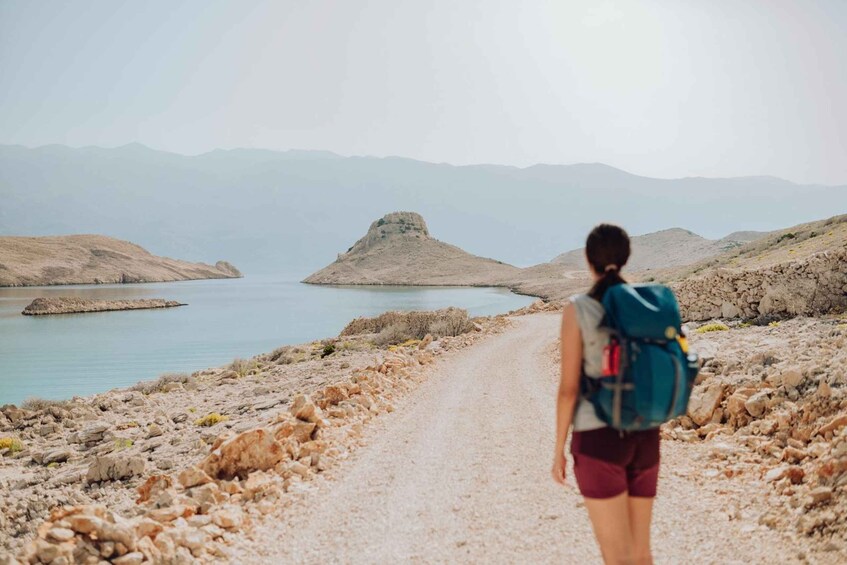 A hiker with a backpack walking on a rocky path beside a body of water with mountains in the background. 1