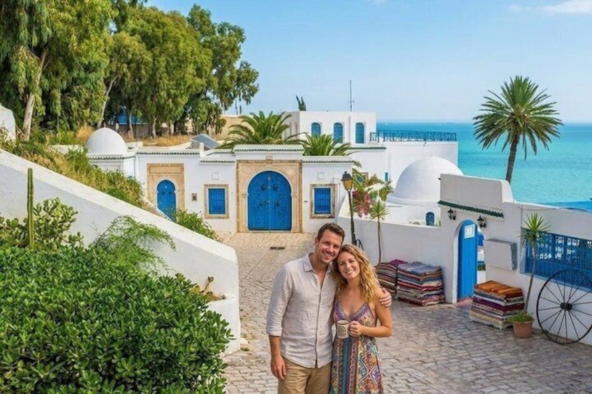 Un couple se tient sur un chemin pavé de pierres, avec en arrière-plan des bâtiments blancs et des portes bleues, d’où l’on aperçoit la mer. 2