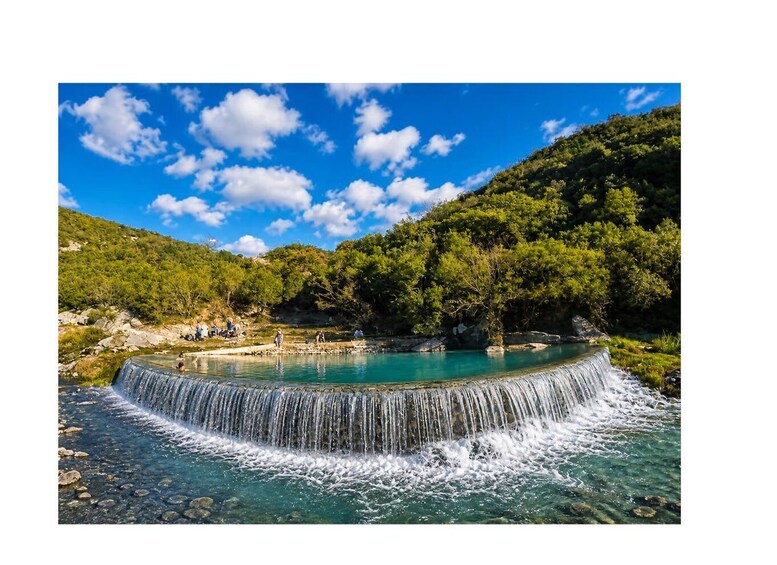 A waterfall cascading into a clear pool surrounded by lush greenery and a forested hillside. 1