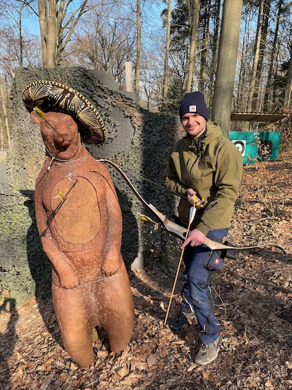 A person with a bow and arrow, standing next to a large bear sculpture in a forest. 4