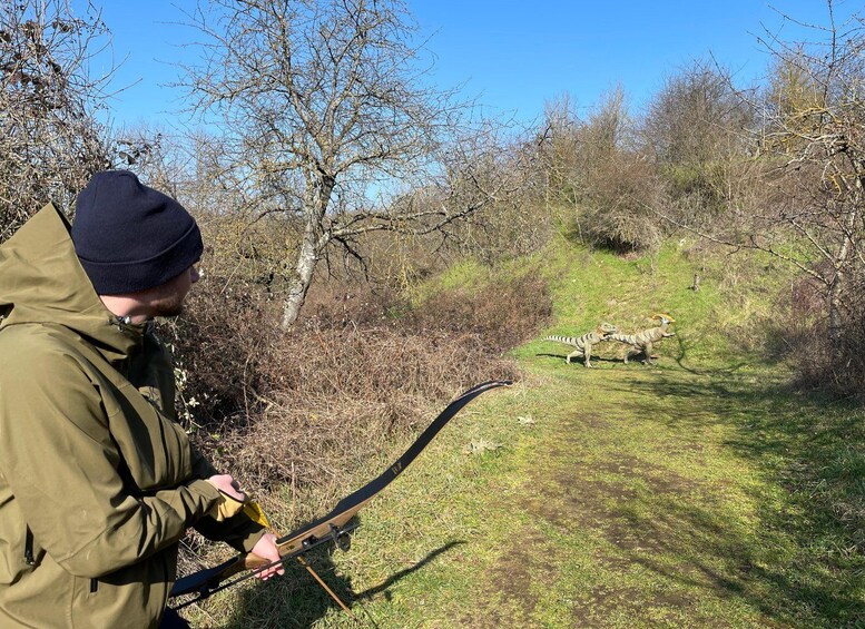 A person in a green jacket and beanie holding a bow, aiming at two camouflaged animals in a wooded area. 5
