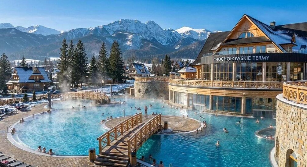 A hot spring resort with a large pool, wooden walkways, and a mountain backdrop. 1