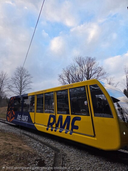 A yellow tram with 'RMF' and 'www.rmt.pl' on its side, parked on tracks with trees and a cloudy sky in the background. 3