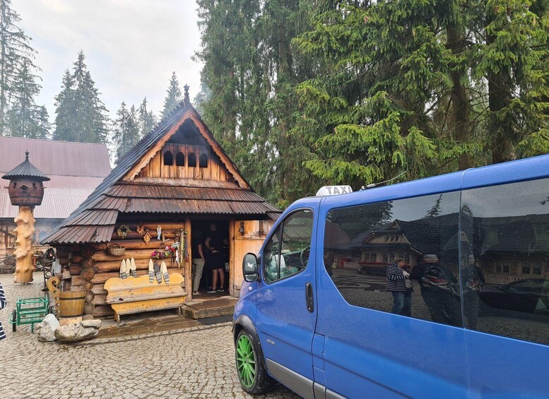 A rustic wooden building with a thatched roof, a blue van parked in front, and a forested backdrop. 2
