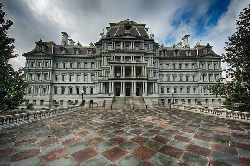A large, symmetrical building with a central entrance, surrounded by a paved plaza and bordered by trees. 5