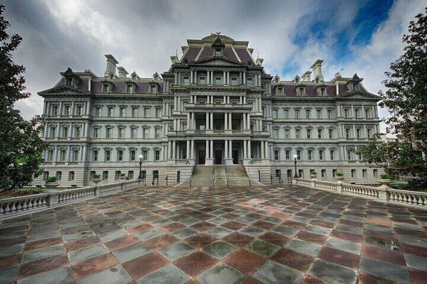 A large, historic building with a symmetrical facade, a central entrance, and a paved courtyard. 3