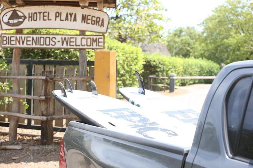 Et træskilt med teksten 'Hotel Playa Negra' og 'Bienvenidos-Welcome', en lastbil med en surfbræt på ladet og et træhegn. 5