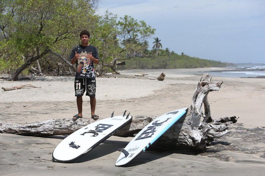 En person står på en sandstrand med to surfboards lænet op ad en drivtømmerstamme. 2