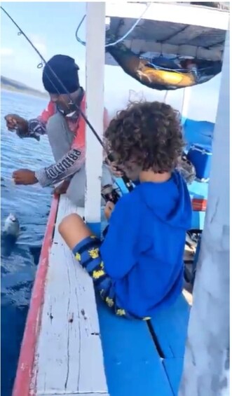 A person in a blue hoodie on a boat, holding a fishing rod. 3