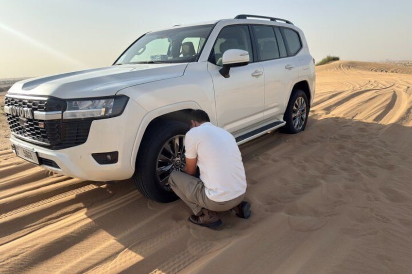 A white SUV parked in a desert with a person crouching near the front wheel. 5