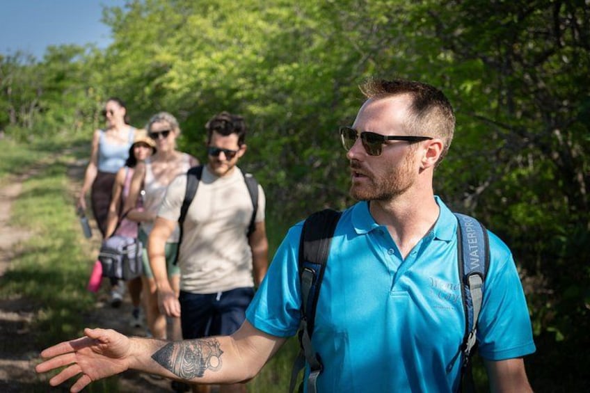 Una persona con un tatuaje en el brazo, con una camiseta azul y una mochila, caminando por un sendero con vegetación al fondo. 1