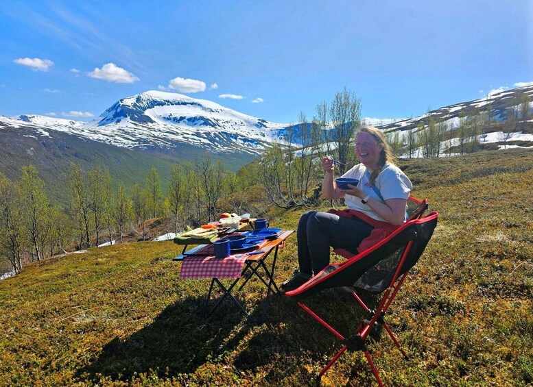 A person sitting on a red chair with a picnic setup on a grassy hill, overlooking a snow-capped mountain. 5