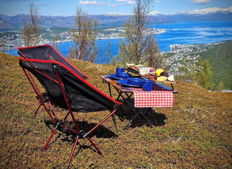 A camping setup with a red and black chair, a table with blue dishes, and a red and white checkered tablecloth on a grassy hill overlooking a lake and mountains. 2