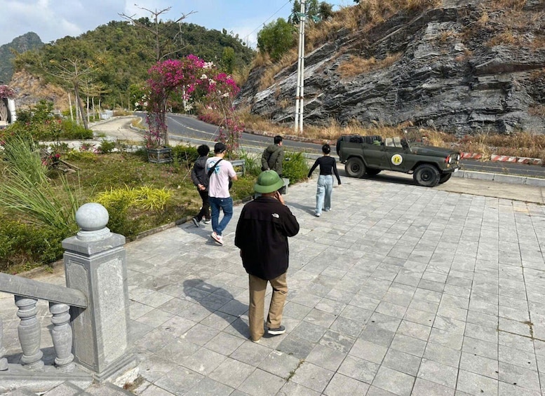 A paved walkway with people walking, a green vehicle, and a rocky hillside in the background. 3
