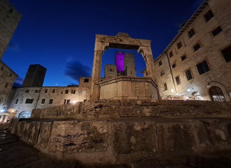 An ancient stone structure with a purple-lit tower, surrounded by old buildings at dusk. 2