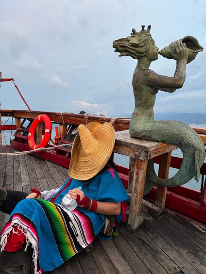 A person sitting on a wooden deck with a straw hat, a colorful blanket, and a statue of a creature with a horned head. 2