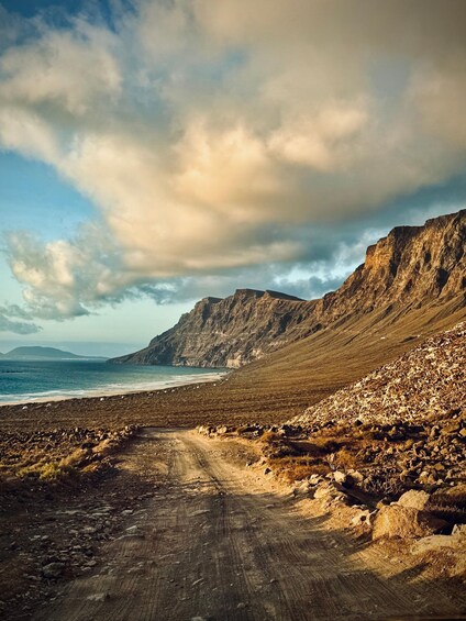 A rugged coastal landscape with a dirt path leading to the beach, rocky terrain, and cliffs under a partly cloudy sky. 1