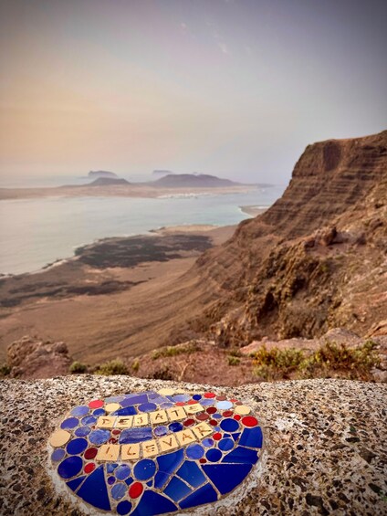 A mosaic on a rocky surface with the word 'DEJATE' and 'LEVAD' in the foreground, overlooking a coastal landscape with cliffs and the sea. 2