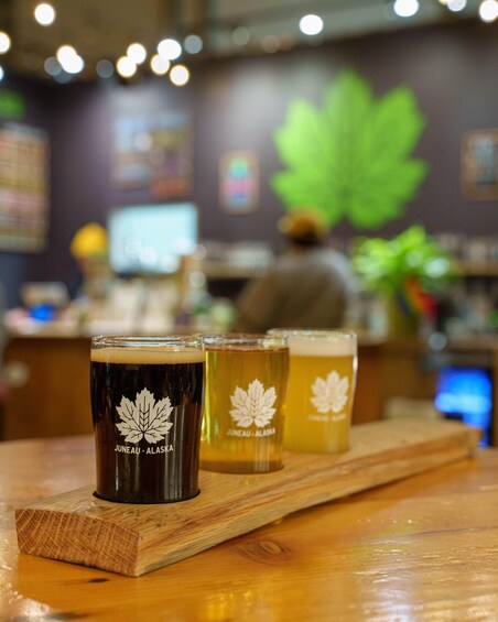 Three glasses of beer on a wooden tray with a maple leaf logo, in a bar with a person in the background. 1