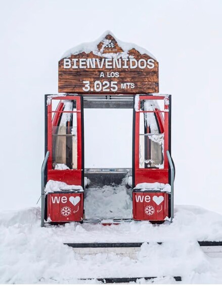 A red vehicle with open doors and a wooden sign above reading 'BIENVENIDOS A LOS 3.025 MTS' with snow on the ground. 1